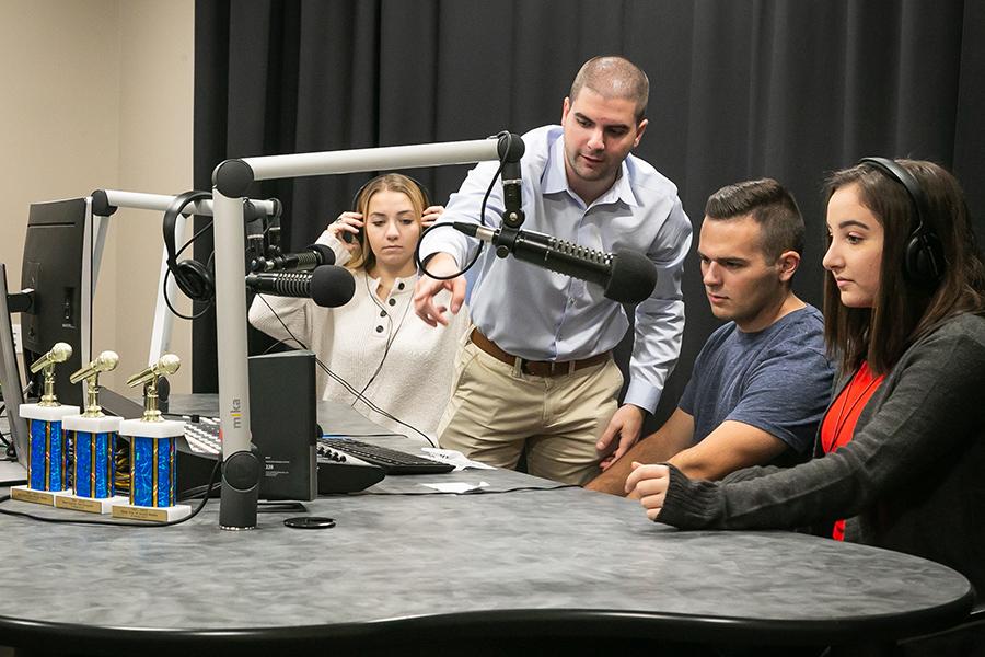 Three students sit around the podcast station, complete with soundboard and microphones, as their professor explains how to use the equipment.