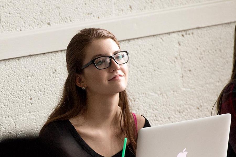 A student with a pony tail and glasses smiles and looks up toward the front of the room during class.