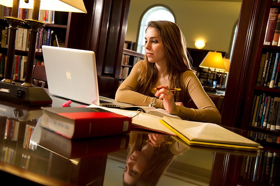 A student in the library works on her mac in the library and consults her written notes.