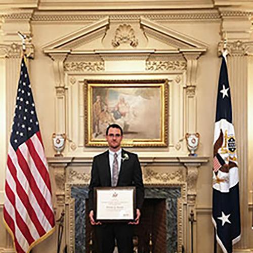 Nicholas Barnett posing with his Edward R. Murrow award, flanked with an American flag on his right and a Washington D.C. flag on his left.