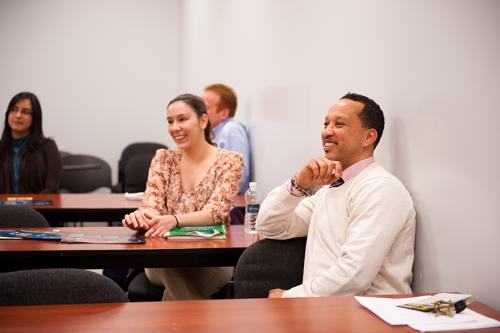 Graduate students work in a classroom and one wears a white sweater with a tie.