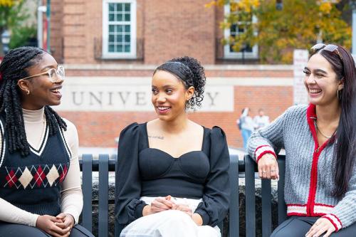 Three students sit on a bench near the North Ave. entrance to Iona.