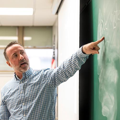 A teacher points to writing on a blackboard.