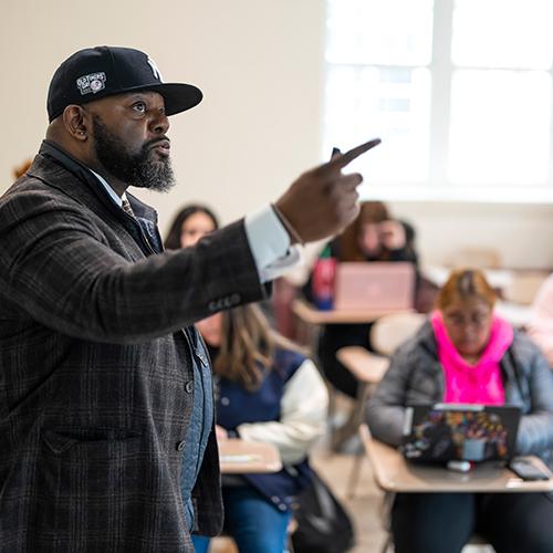 A teacher lectures in front of a class in a Yankees cap and blazer.